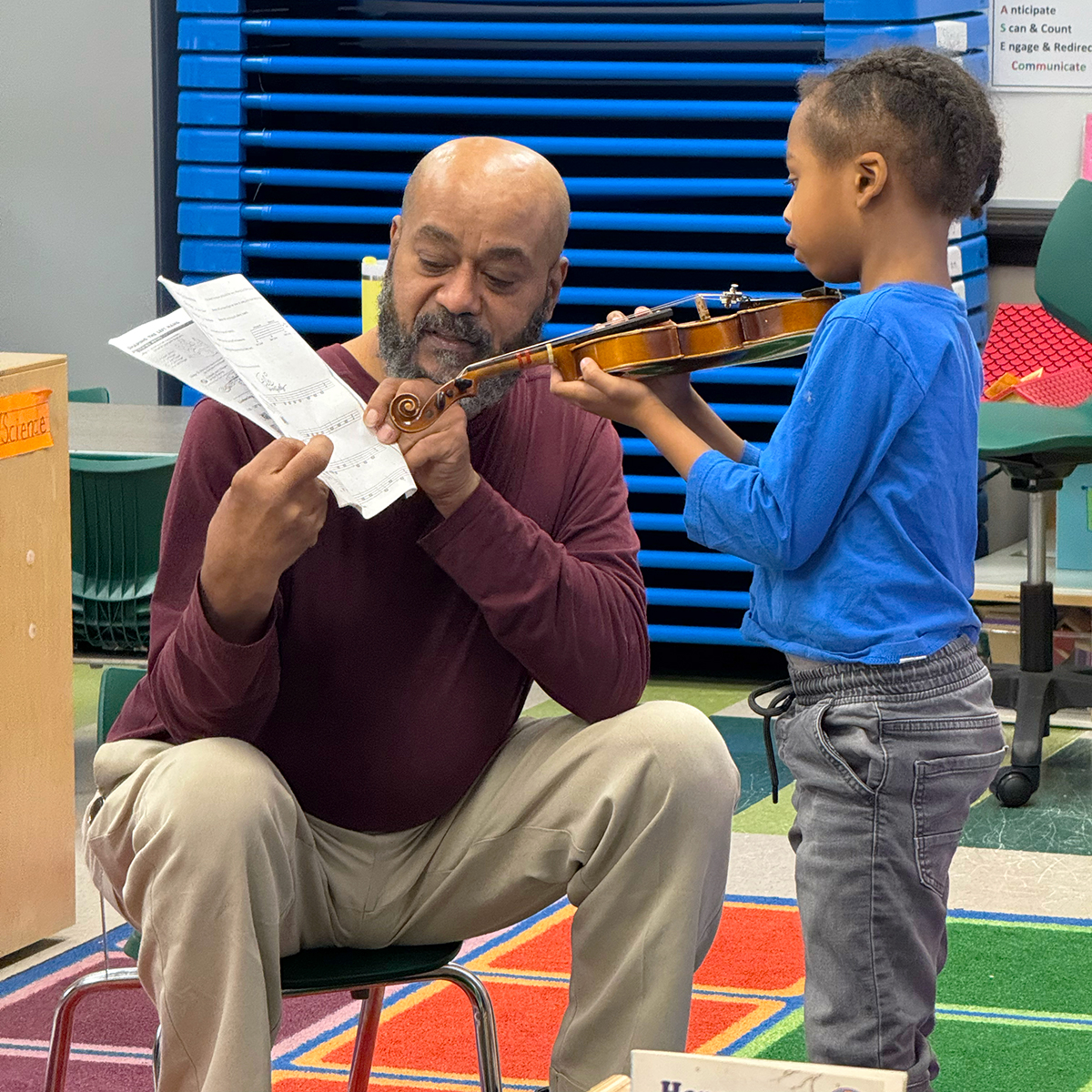 Learning Through Music A young student plays the violin while a teacher sits and points to sheet music
